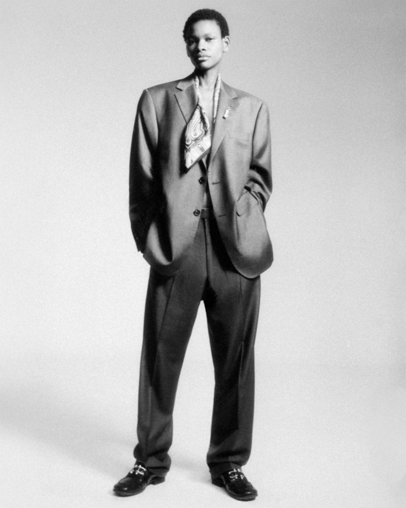 Model standing in an oversized suit and silk scarf against a plain studio background