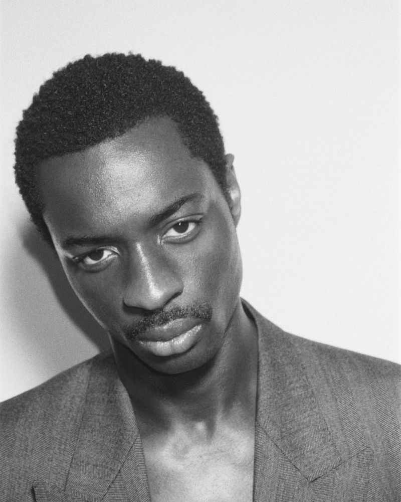Close-up monochrome portrait of a Black man wearing a tailored suit jacket against a plain white wall.