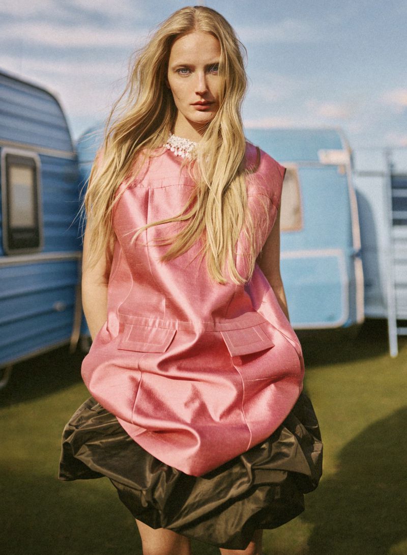 Blonde woman wearing a pink sleeveless dress and dark skirt standing outdoors in front of a blue caravan for Elle Greece.