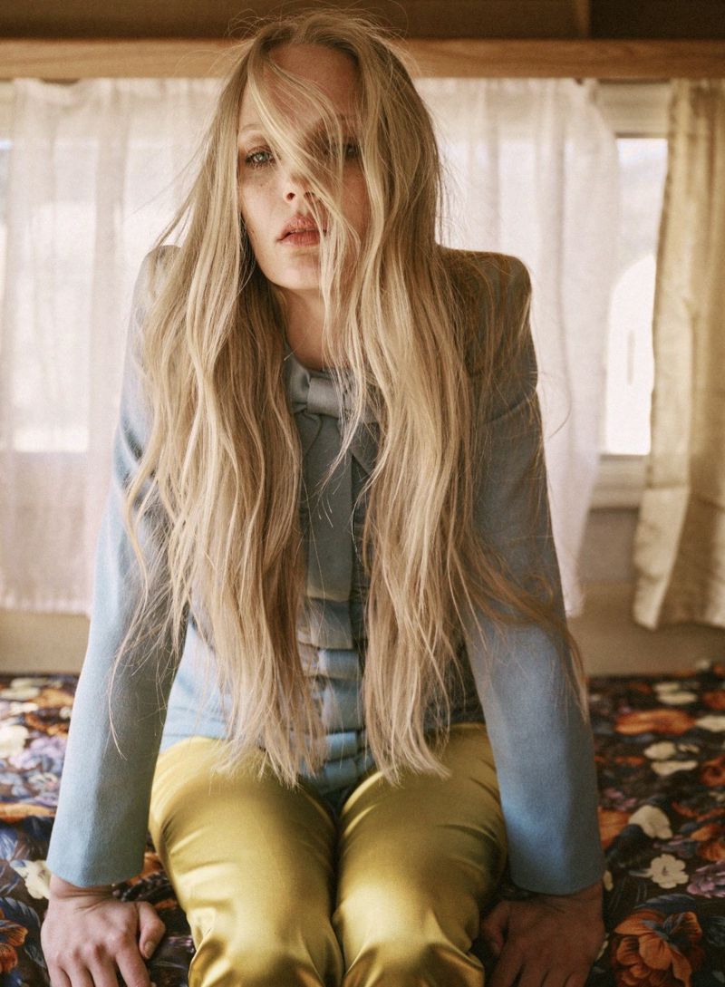 Blonde woman wearing a blue jacket and yellow trousers inside an interior space with floral bedding and white curtains.