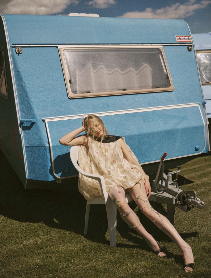 Blonde woman seated in a white plastic chair against a vintage blue caravan for an Elle Greece fashion editorial.