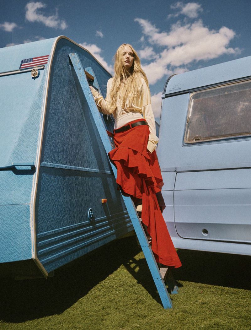 Blonde woman in a yellow top and red ruffle skirt standing on a ladder between two blue trailers under a cloudy sky.
