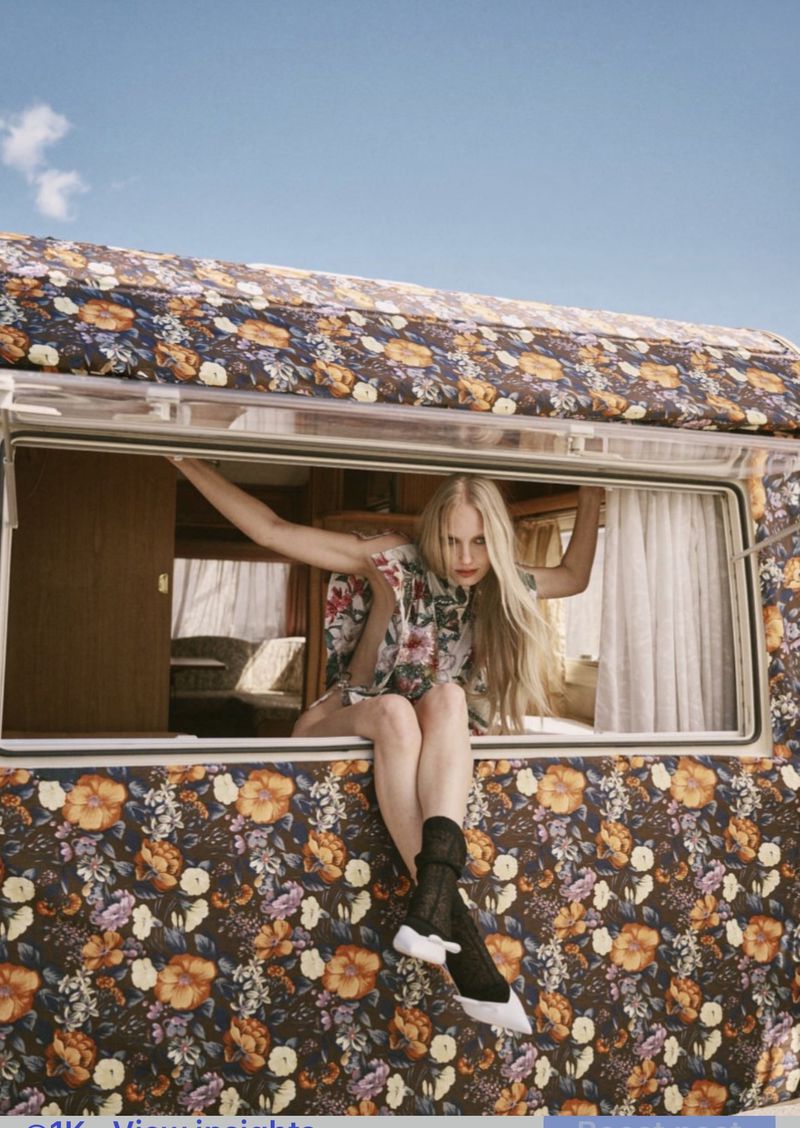 Model in floral print top sitting in the window of a patterned camper van under a blue sky