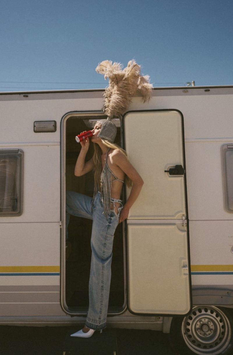 Woman in feathered headpiece and denim jeans standing in a camper van doorway drinking from a can against a clear blue sky