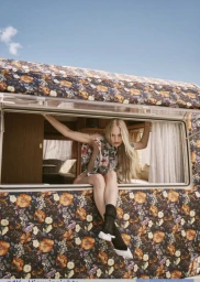 Model in floral print top sitting in the window of a patterned camper van under a blue sky