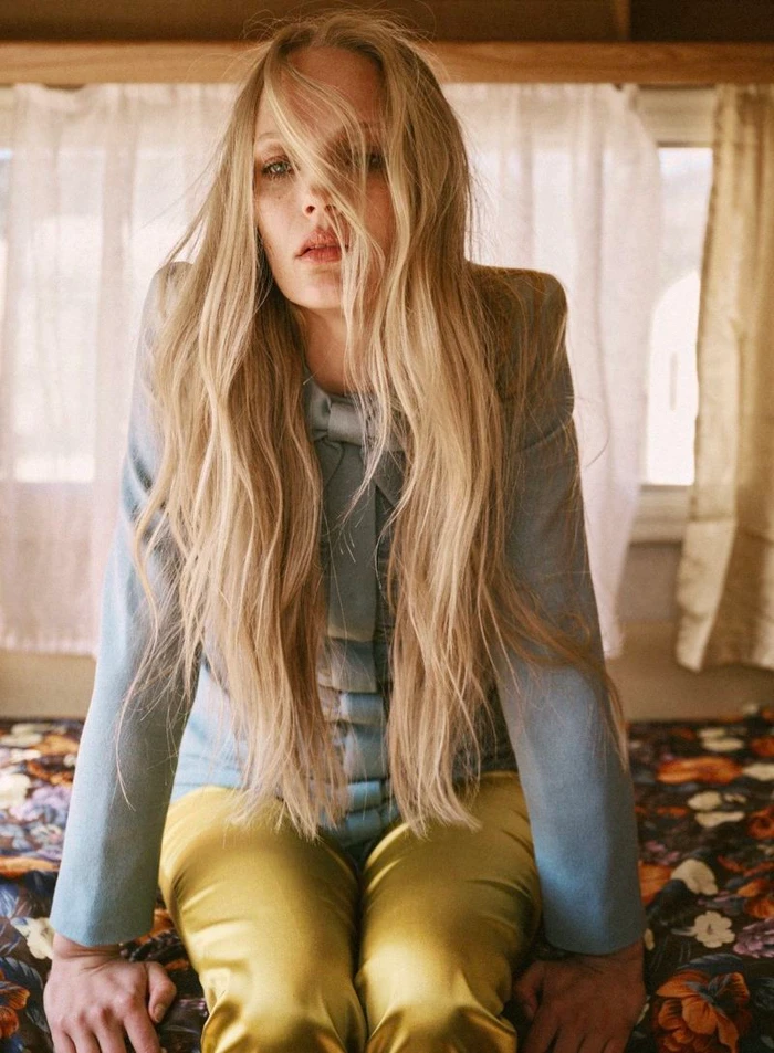 Blonde woman wearing a blue jacket and yellow trousers inside an interior space with floral bedding and white curtains.
