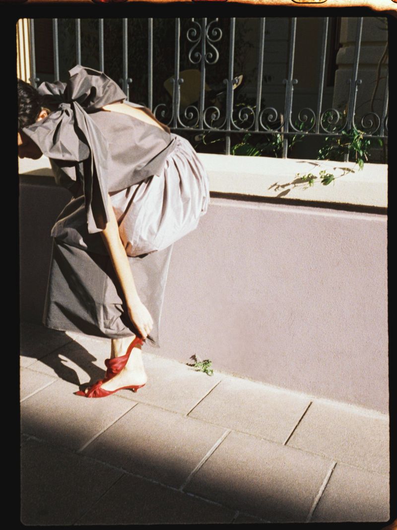 Woman in a gray dress and red shoes adjusts her sandal strap in an outdoor setting next to a wall and black wrought-iron fence.
