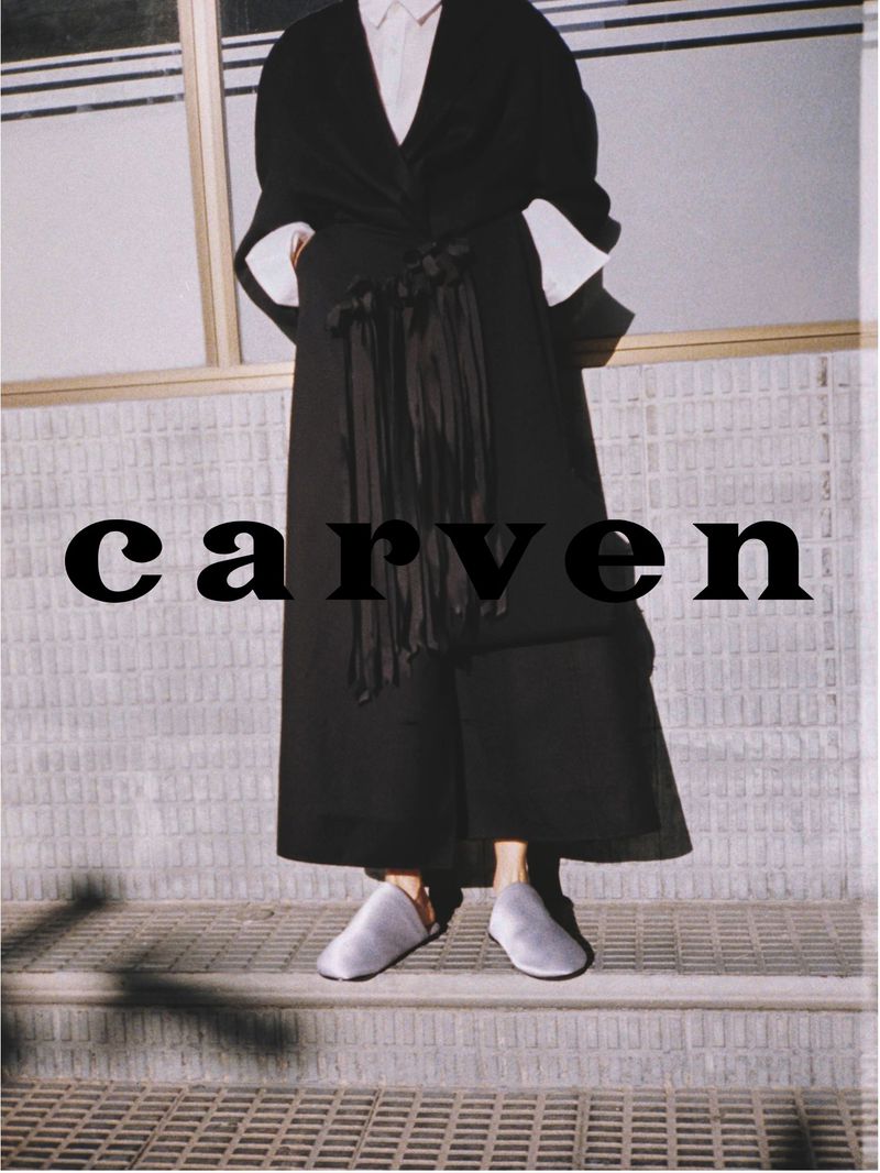 Fashion shot of an adult female model wearing a long black coat, white shirt, and gray shoes. She stands on a concrete step outside.