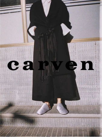 Fashion shot of an adult female model wearing a long black coat, white shirt, and gray shoes. She stands on a concrete step outside.