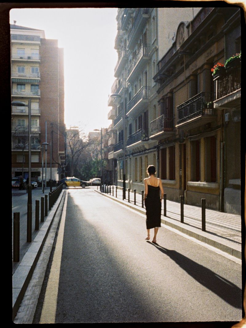 Woman in black dress walks down an empty city street on a sunny day, flanked by tall buildings, creating a serene and fashionable scene.