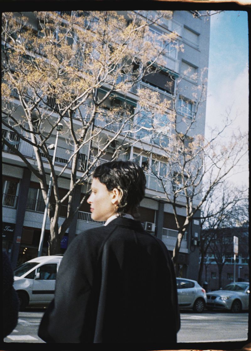 Adult woman walks along a city street near bare trees and apartment buildings. She wears a dark coat and her head is turned in profile.
