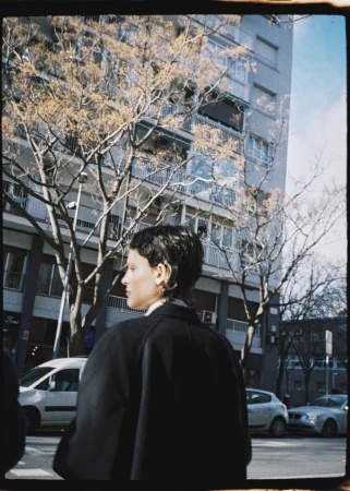 Adult woman walks along a city street near bare trees and apartment buildings. She wears a dark coat and her head is turned in profile.