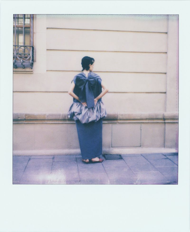 Back view of a woman in a blue dress with a large bow, standing on a sidewalk in front of a beige building with a window.