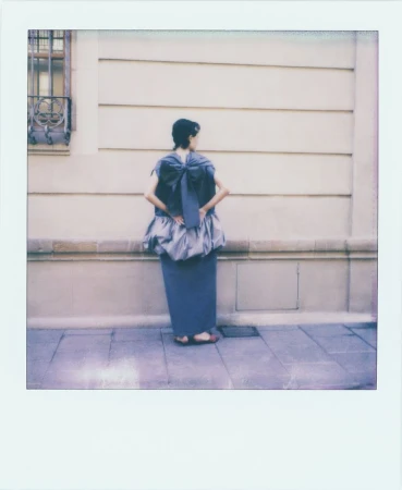 Back view of a woman in a blue dress with a large bow, standing on a sidewalk in front of a beige building with a window.