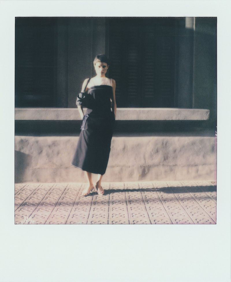 Woman in a black dress stands on a patterned sidewalk in front of a building with shuttered windows on a sunny day.
