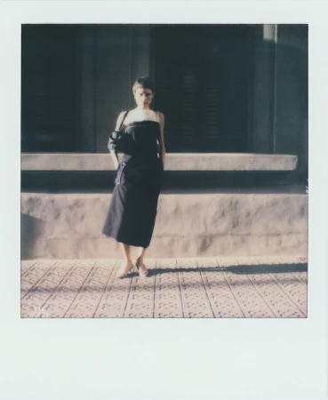 Woman in a black dress stands on a patterned sidewalk in front of a building with shuttered windows on a sunny day.