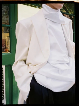 Fashion shot featuring a person in a white blazer and draped white shirt against a green door in diffused lighting.