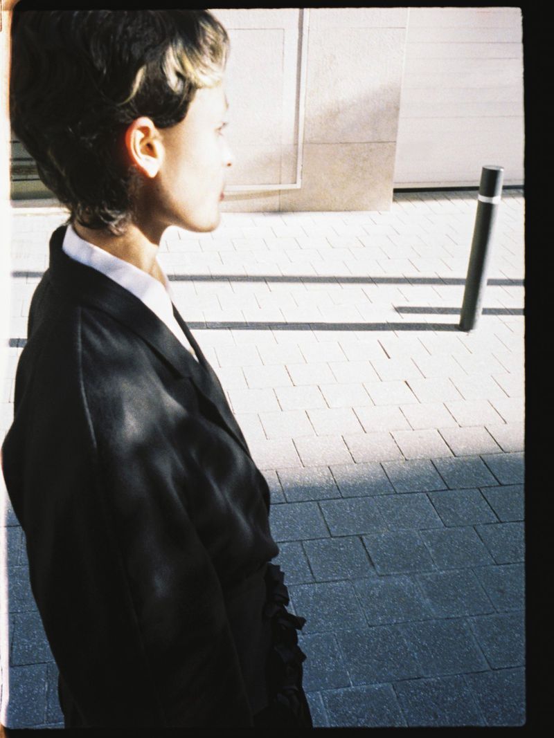 Profile shot of a woman in a suit outdoors on a patterned brick sidewalk. Strong light and shadows highlight the architectural space.
