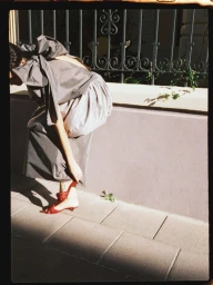 Model wearing a grey bow-back dress adjusting red heeled shoes by a wall with iron railings.