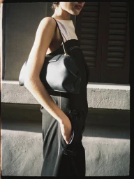 Model wearing a dark dress holding a leather handbag outdoors against an architectural background