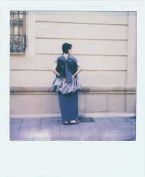 Polaroid photograph of a model seen from behind wearing a grey dress with a large bow against a building wall