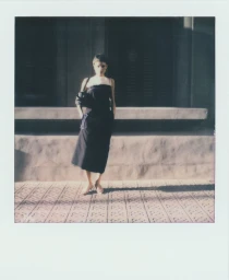 Model wearing a dark dress standing on a tiled sidewalk in front of a building exterior during CARVEN S/S 25