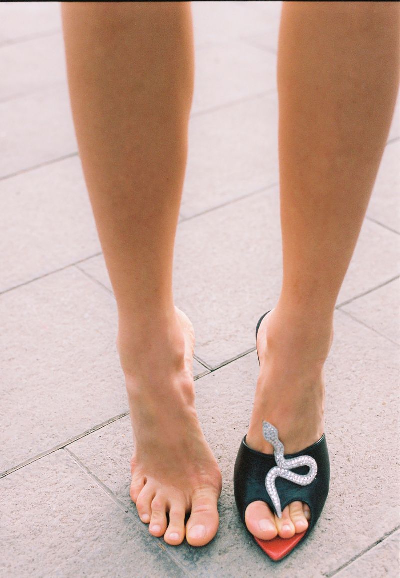 Close-up shot of bare legs and feet standing on stone paving, one foot wearing a black mule shoe with a crystal snake detail