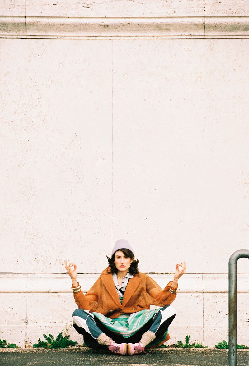 Model sitting cross-legged in a meditative pose against a pale stone wall wearing an orange coat and beanie