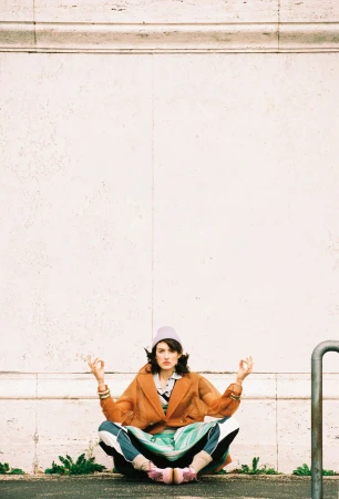 Model sitting cross-legged in a meditative pose against a pale stone wall wearing an orange coat and beanie