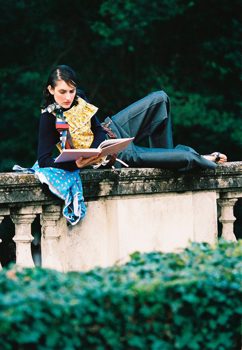 Woman in wide-leg pants and a yellow top sitting on a stone wall, reading a book in a garden with green foliage.