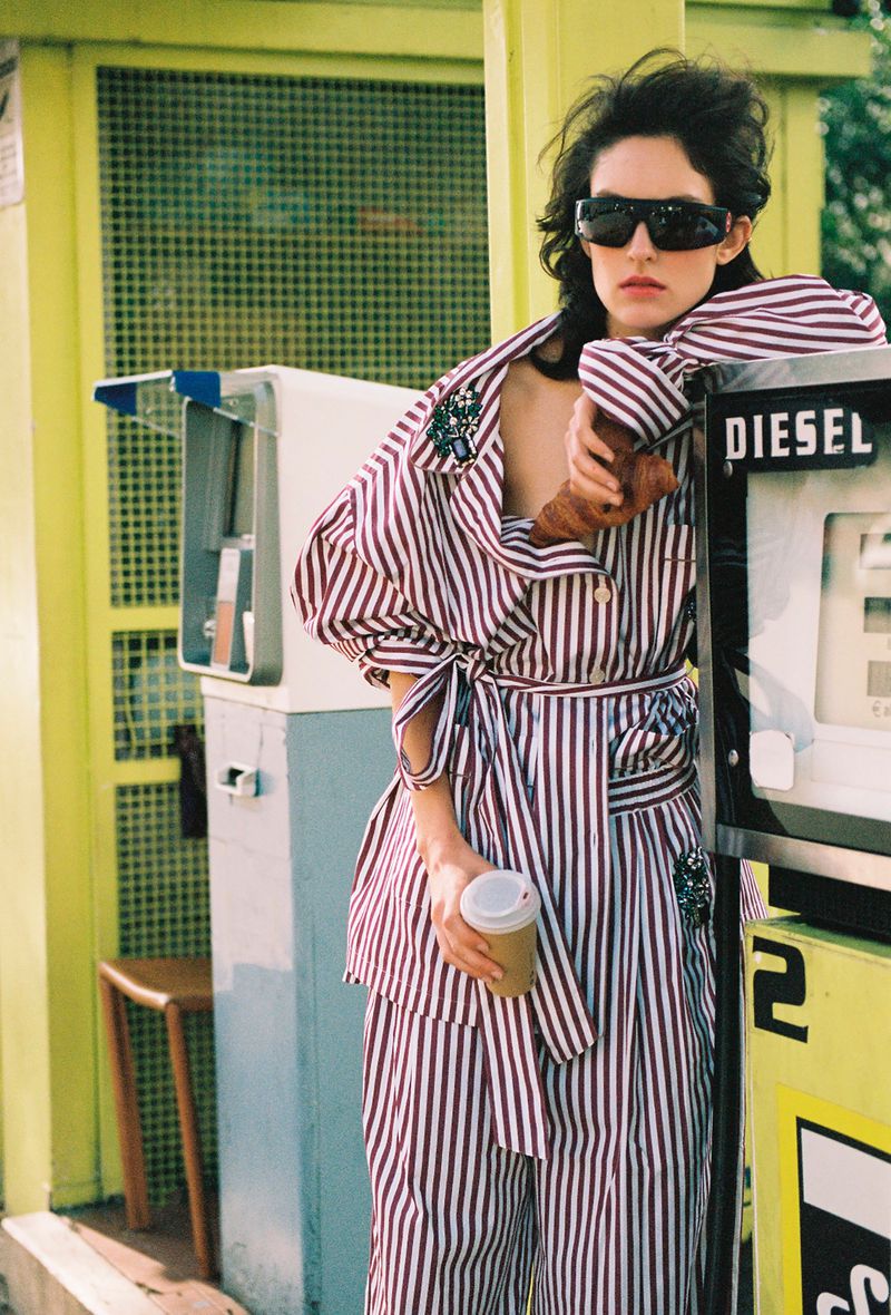 Model in striped shirt and matching trousers leaning against a petrol pump holding a paper cup and a croissant