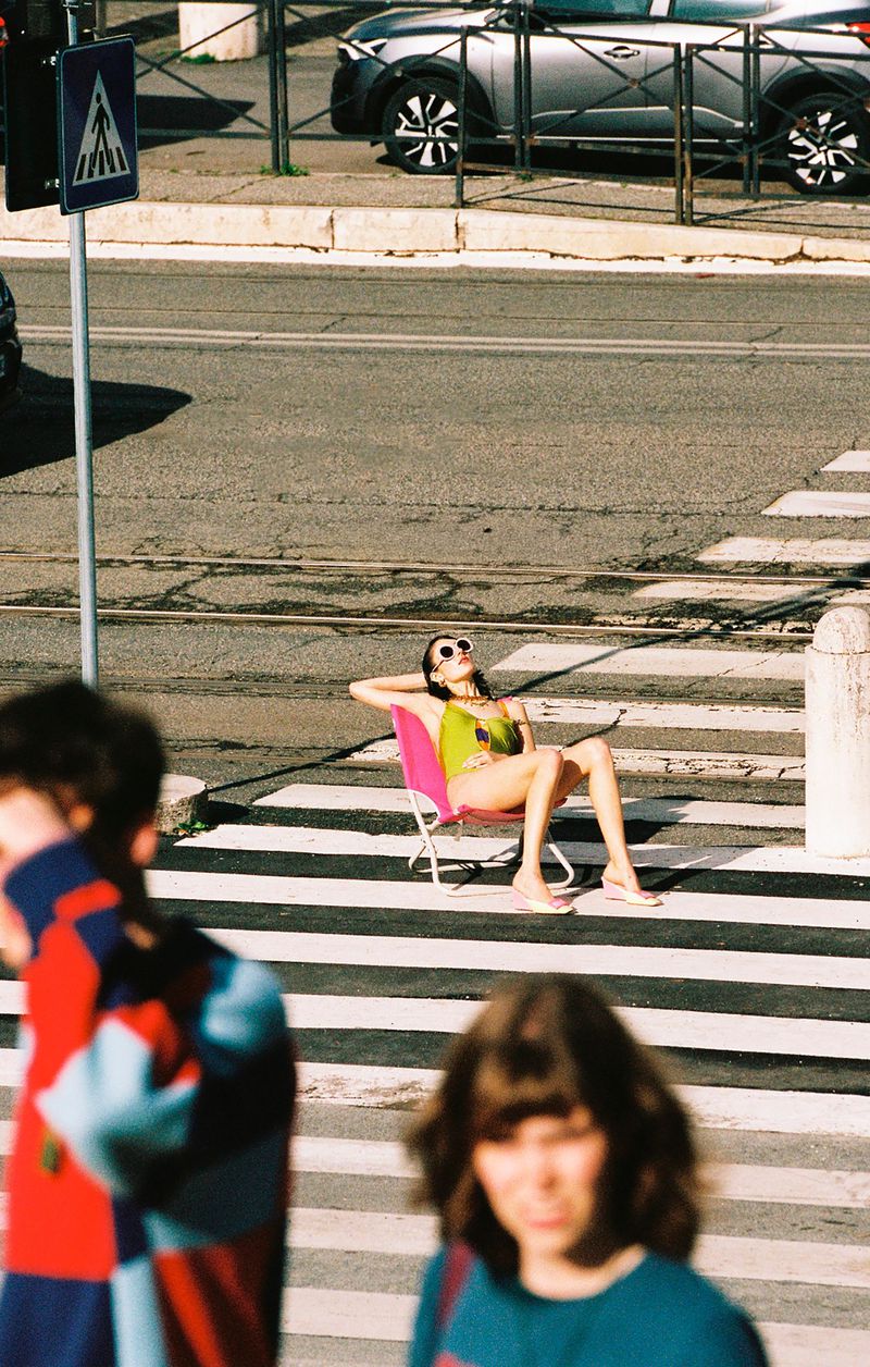 Model relaxing on a pink folding chair in the middle of a street crosswalk with out-of-focus figures in the foreground
