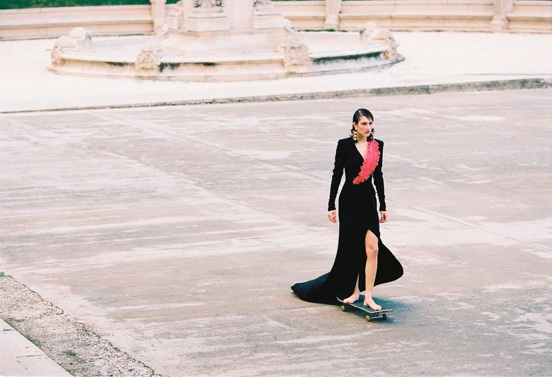 Model in a black gown with pink detailing riding a skateboard across an open paved public square
