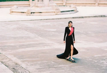 Model in a black gown with pink detailing riding a skateboard across an open paved public square