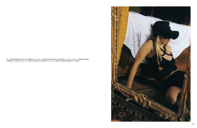 Model seated on the floor leaning against a bed, wearing a black top, dark skirt, and a large black hat.