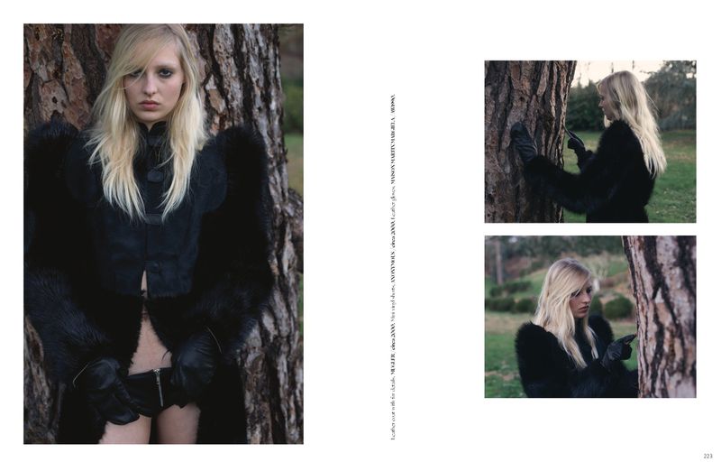 Editorial portrait of a blonde woman wearing black outerwear and leather gloves posing by a tree trunk for a magazine layout.