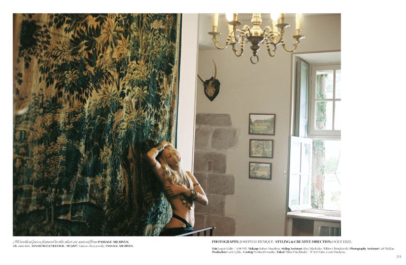 Model posing against a large ornate tapestry in an interior room with a rustic stone wall, chandelier, and open window
