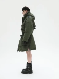 Model in green puffer coat wearing dark beanie and black boots against a plain white background
