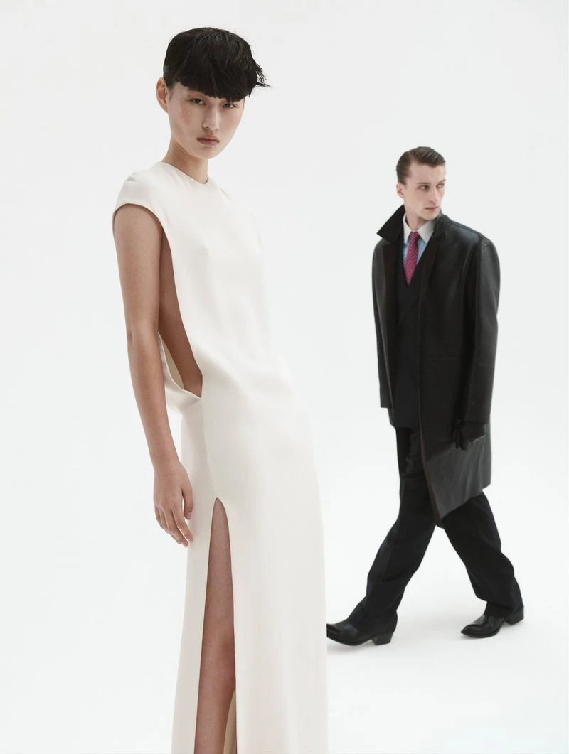 Female model in a long white sleeveless dress in the foreground, with a male model in a dark coat behind her.