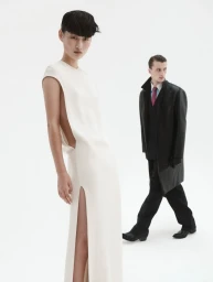 Female model in a long white sleeveless dress in the foreground, with a male model in a dark coat behind her.