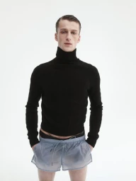 Young man in a black turtleneck sweater and light blue shorts standing against a bright white background.