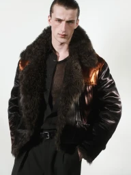 Male model wearing a dark shearling collar bomber jacket and black shirt against a white seamless background
