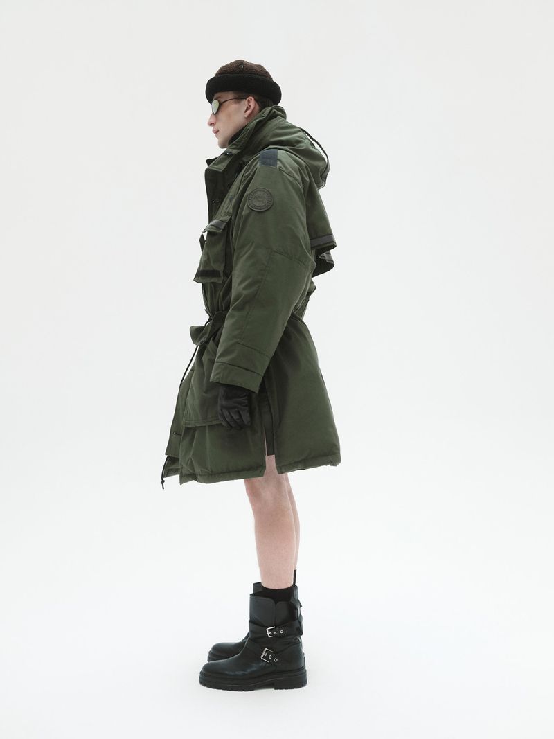 Model in green puffer coat wearing dark beanie and black boots against a plain white background