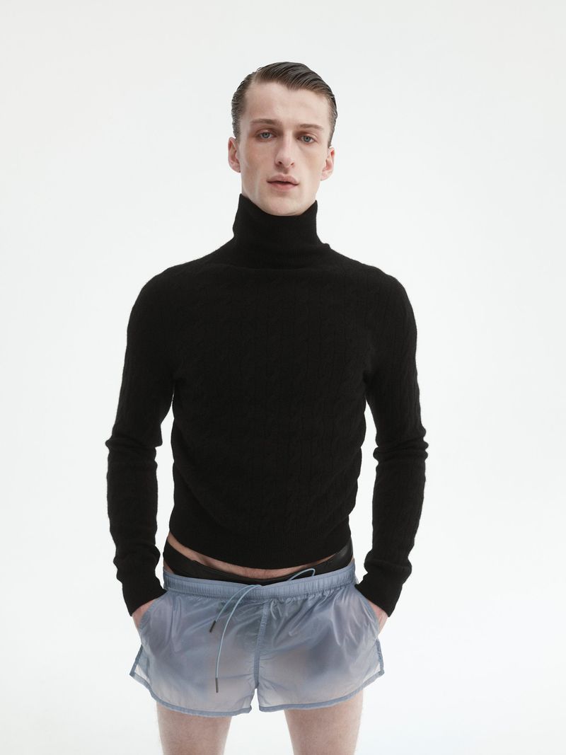Young man in a black turtleneck sweater and light blue shorts standing against a bright white background.
