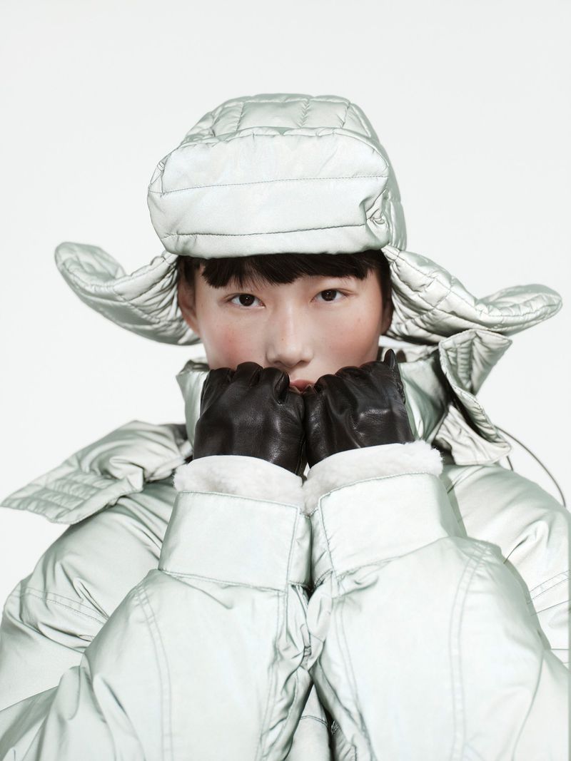 Model wearing a quilted silver puffer hat and jacket holding gloved hands near face