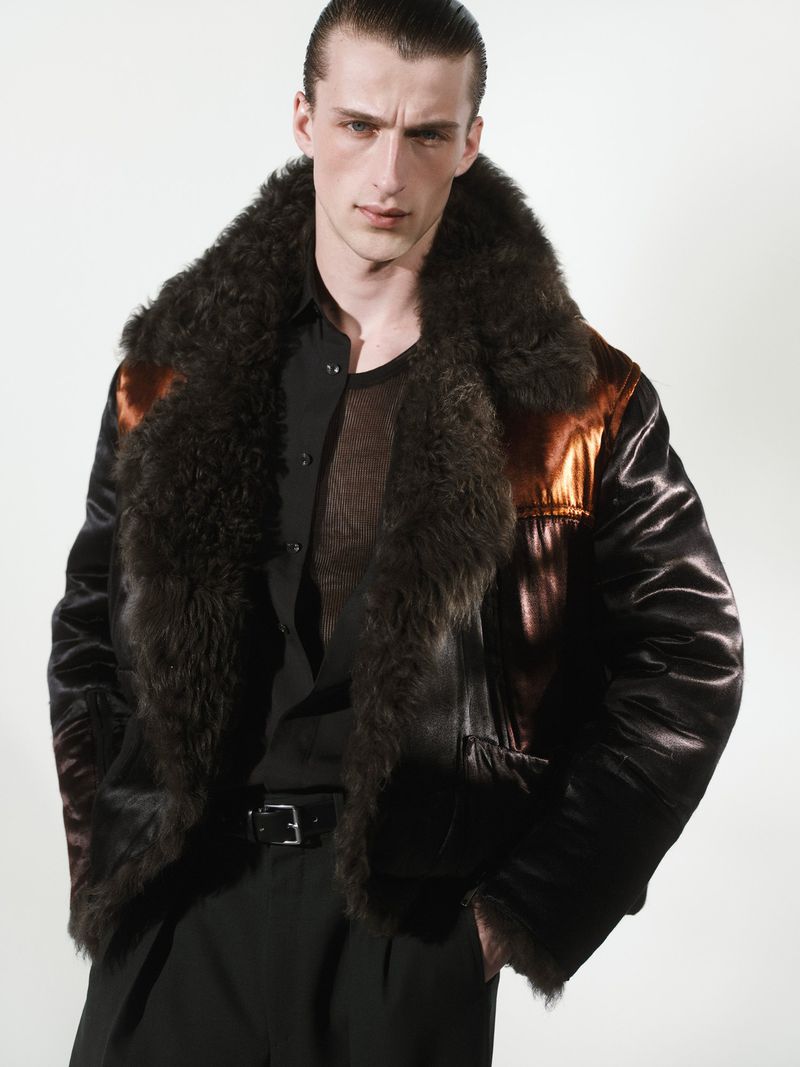 Male model wearing a dark shearling collar bomber jacket and black shirt against a white seamless background