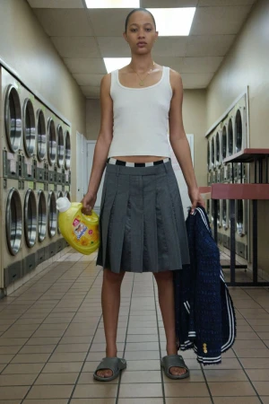 Model wearing a white tank top and grey pleated skirt holding laundry detergent inside a laundromat