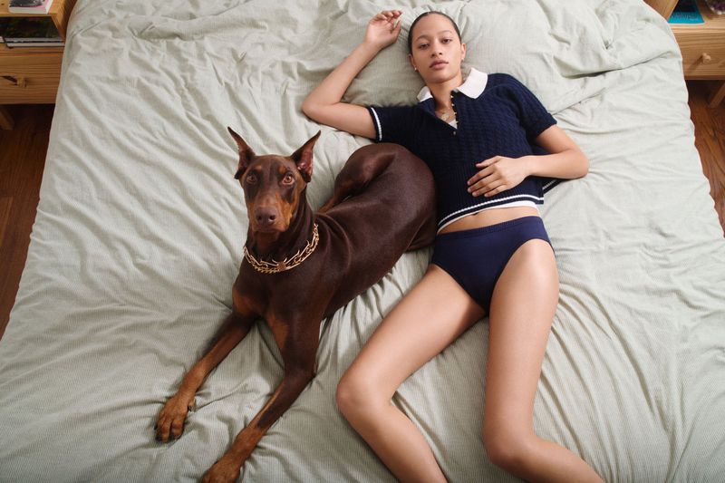 Model Selena Forrest lying on a green bed next to a brown Doberman dog wearing a chain collar for Pop Magazine