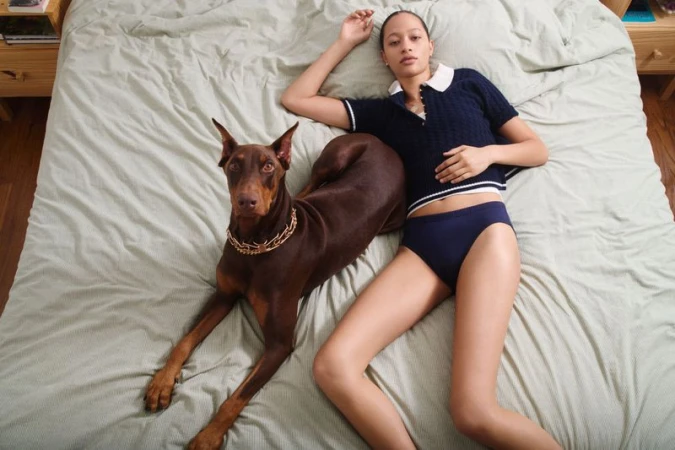 Model Selena Forrest lying on a green bed next to a brown Doberman dog wearing a chain collar for Pop Magazine