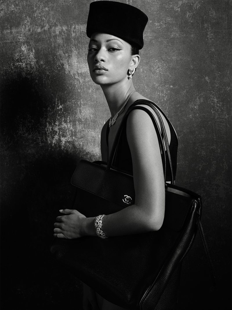 Portrait of an adult woman wearing a hat, jewelry, and a black tank top carrying a large handbag in front of a mottled grey background.